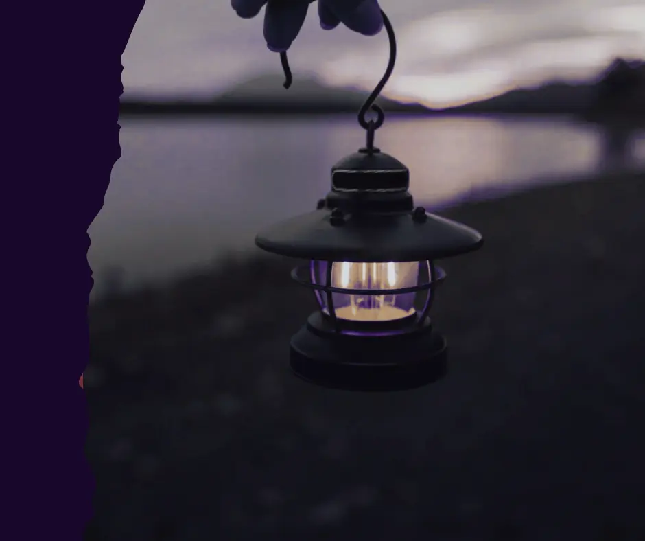 A hand holding a small, glowing black lantern with a warm purple light, against a blurred, dark background of a lake and mountains at dusk.