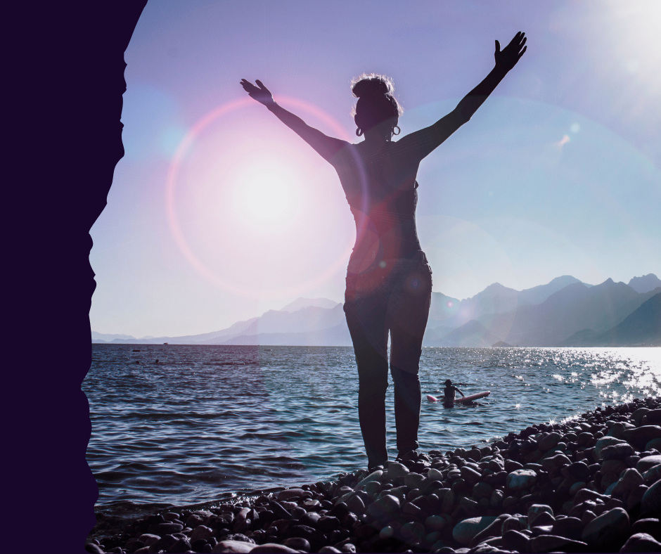 A backlit silhouette of a woman standing on a pebble beach with her arms raised in celebration or surrender, facing the bright sun setting or rising over a calm body of water.