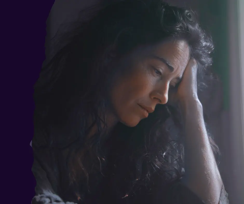 Close-up of a middle-aged woman with dark, wavy hair looking down, leaning her head on her hand, suggesting exhaustion, stress, or deep contemplation due to chronic depletion.