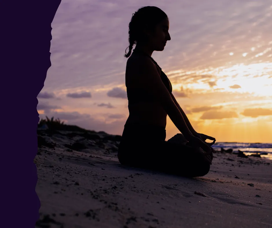 A silhouette of a woman sitting in a meditative yoga pose on a sandy beach at sunset. She is facing the ocean with her hands resting on her knees in a mudra position, captured during the golden hour with soft purple and orange clouds in the background.