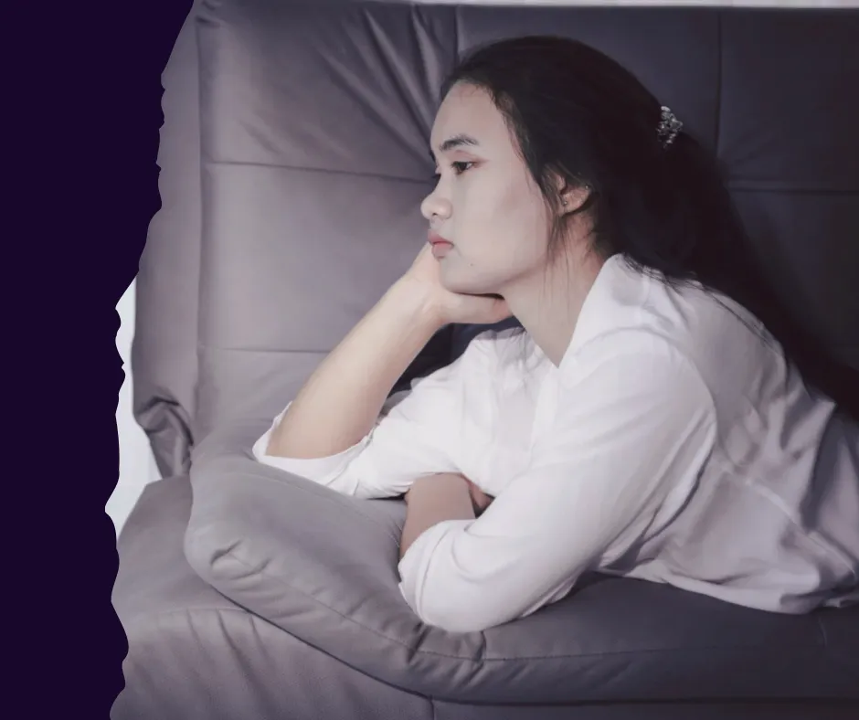 Woman sitting on a couch looking thoughtful and emotionally drained, representing burnout and nervous system dysregulation.